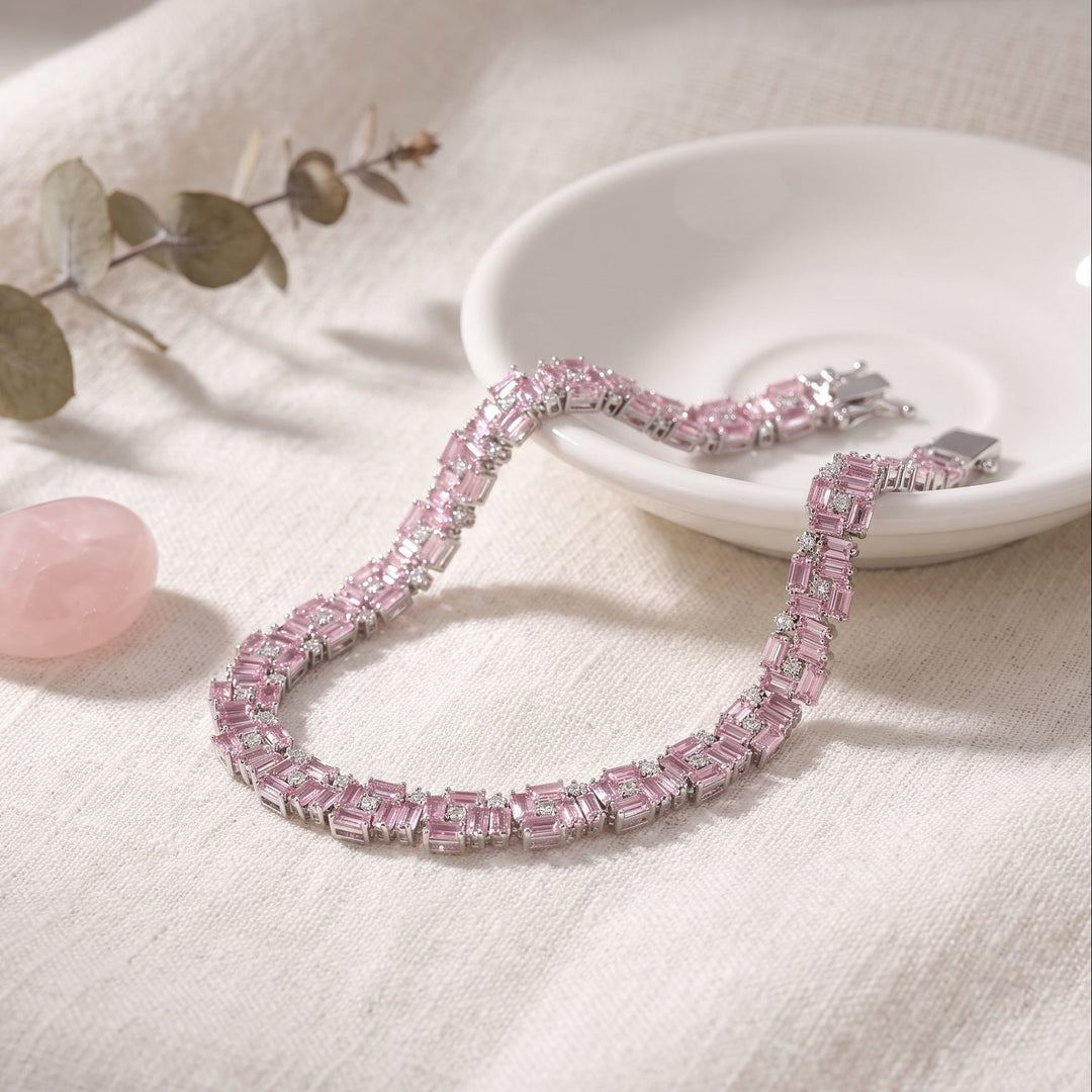 A pink gemstone tennis bracelet with rectangular baguette-cut pink stones in a silver-toned setting, displayed on a textured cream linen surface next to a small white dish, a pink stone, and a eucalyptus sprig.